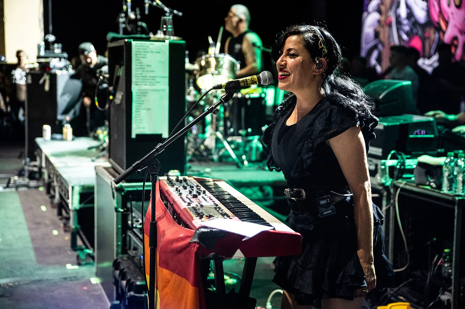 A woman singing into a microphone beside a keyboard on stage, surrounded by musical equipment and band members, in a lively concert setting.
