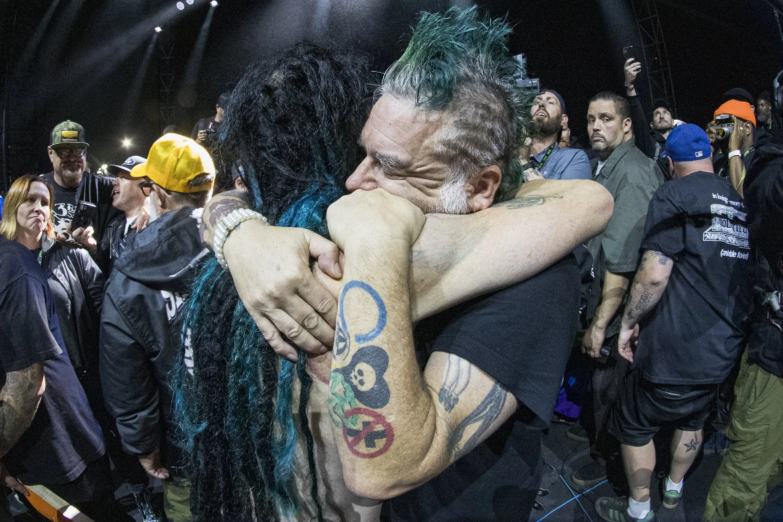 Two people embracing tightly at a concert or event, surrounded by a crowd of onlookers and photographers.