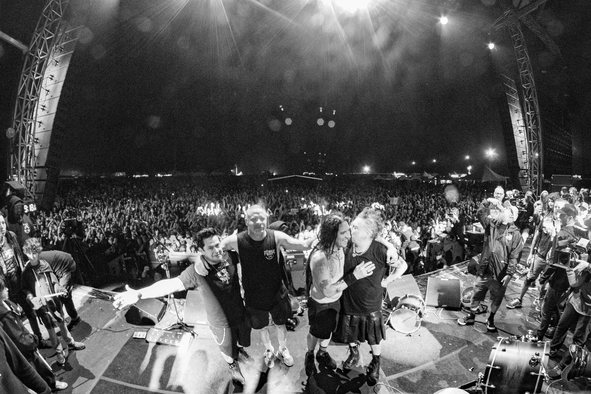 Black and white photo of a concert stage with four performers hugging and smiling, and a crowd of people in the background under a large tent at night.