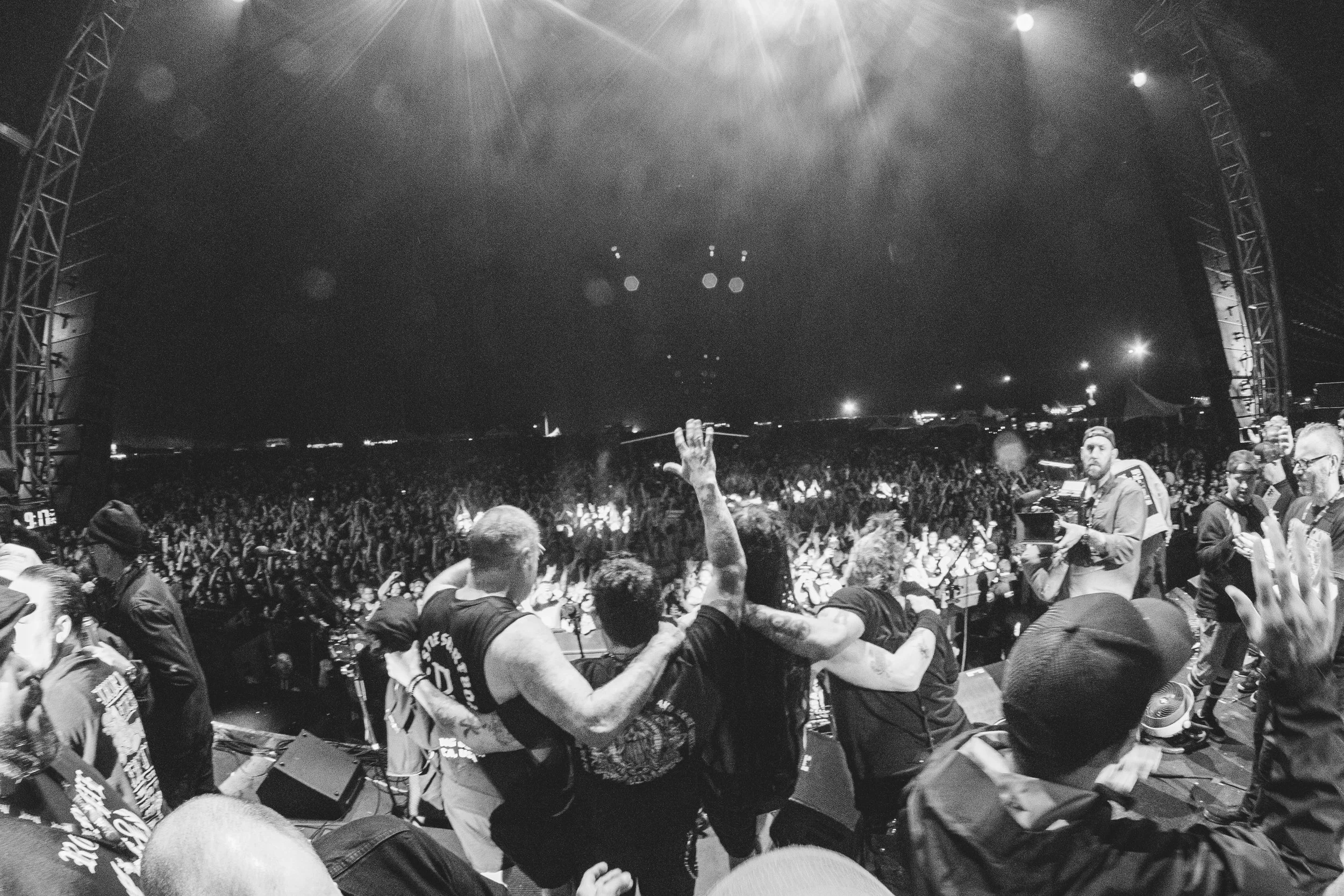 Black and white photo of a concert stage with a large crowd, stage lights, and people on stage and in the audience.