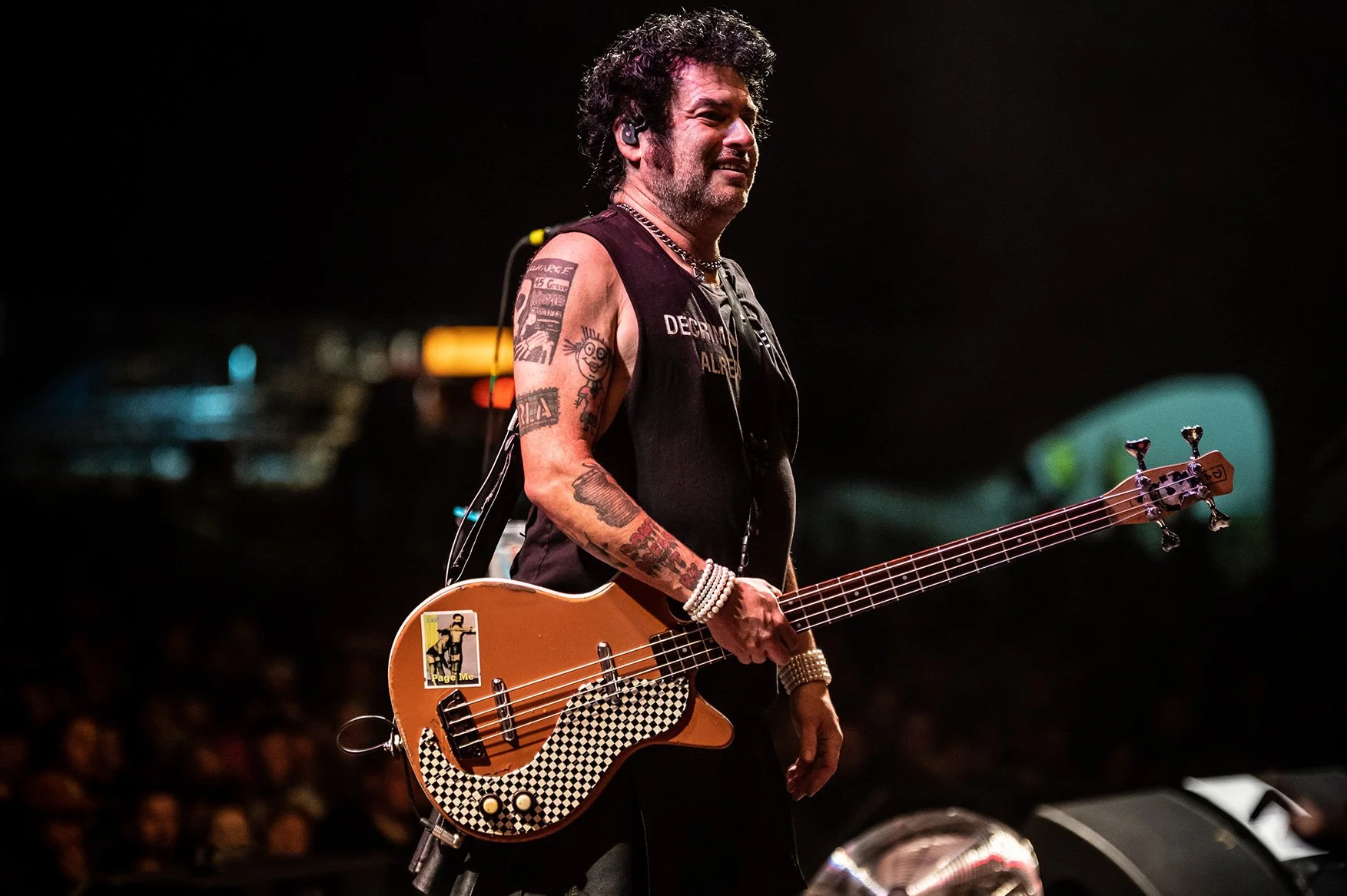A male bass guitarist performing on stage at night, holding a guitar with a checkered pattern on the pickguard. He has curly hair, tattoos on his arms, and is wearing a sleeveless black shirt and jewelry, with a crowd in the background.