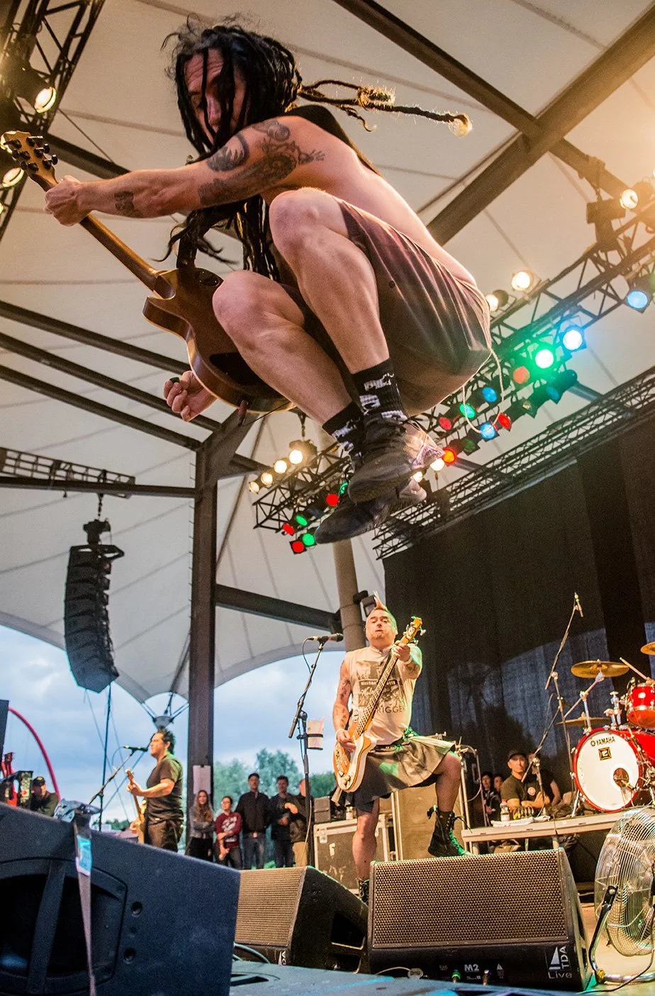 A guitarist jumps mid-air during a live outdoor concert, with band members and spectators visible below.