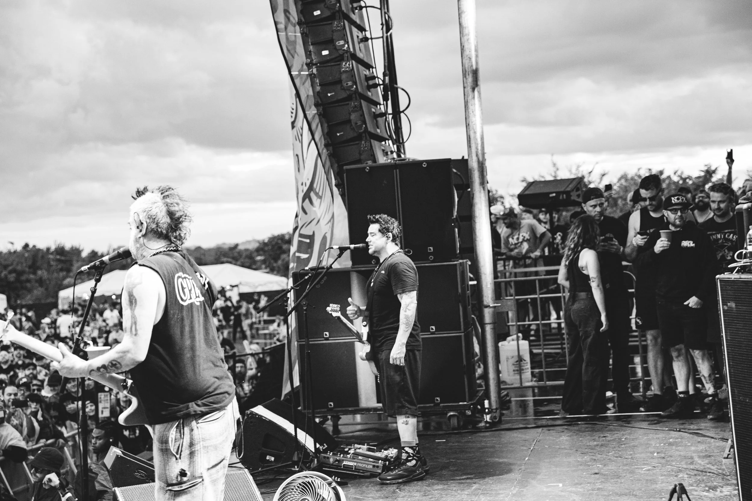 Black and white photo of a live outdoor music concert with a band performing on the stage and an audience in the background.