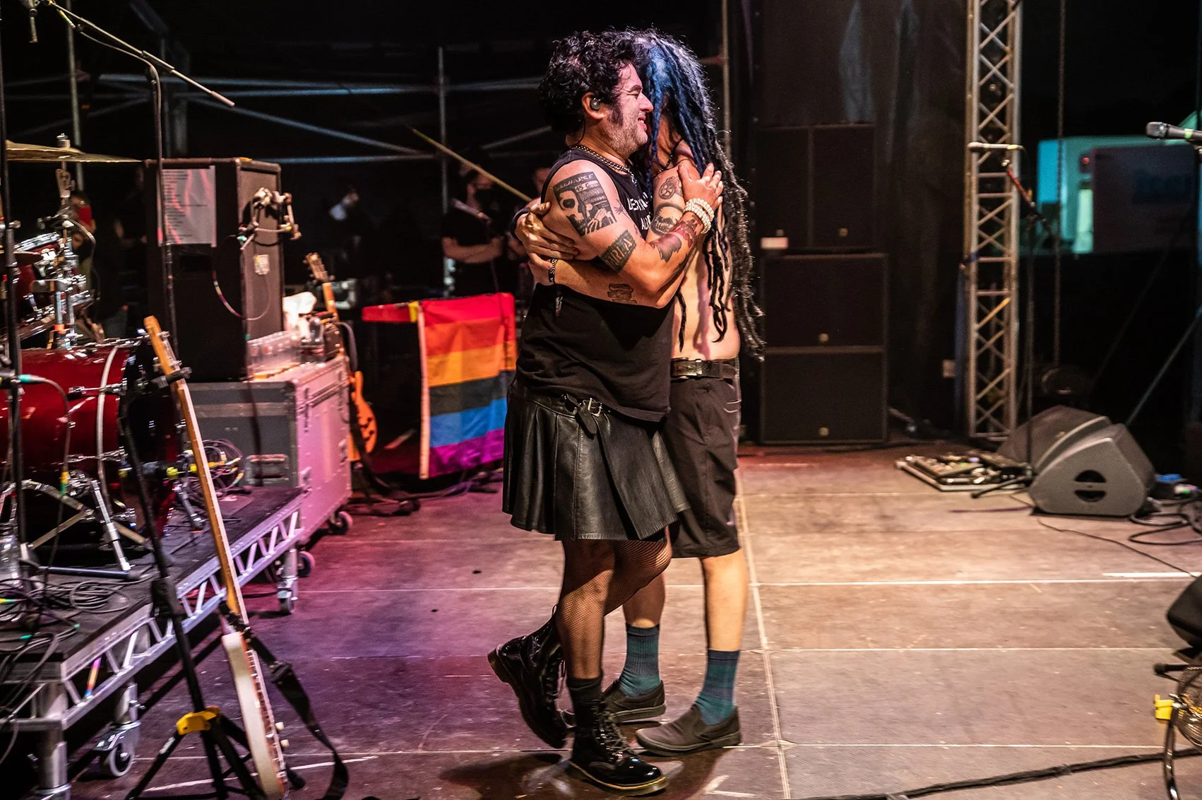 Two people hugging on a stage, one with black hair, tattoos, and wearing a black leather skirt; the other with dreadlocks and shirtless. Stage equipment and a rainbow pride flag in the background.
