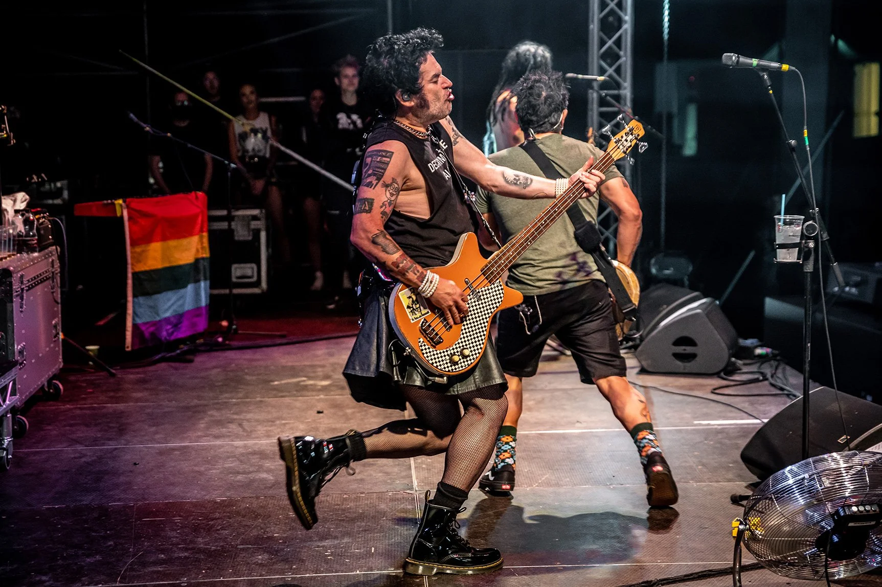 A musician with tattoos, wearing a sleeveless top, skirt, fishnet stockings, and combat boots is playing an electric guitar on stage. The stage has a rainbow flag and various equipment, with audience members visible in the background.