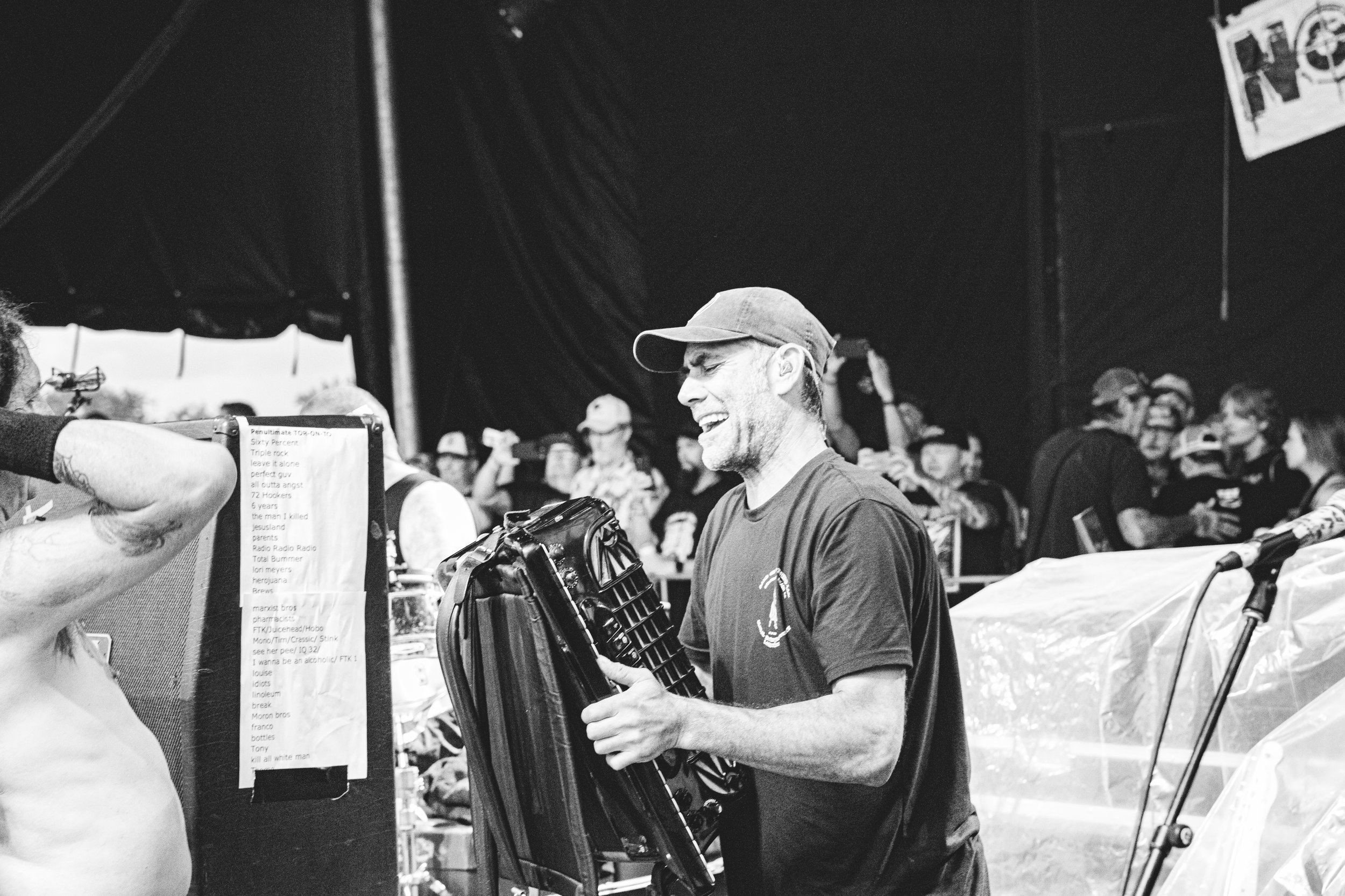 A man with a cap and a T-shirt smiling and laughing while holding a large black object, with a crowd of people behind him at an outdoor event, black and white photo.