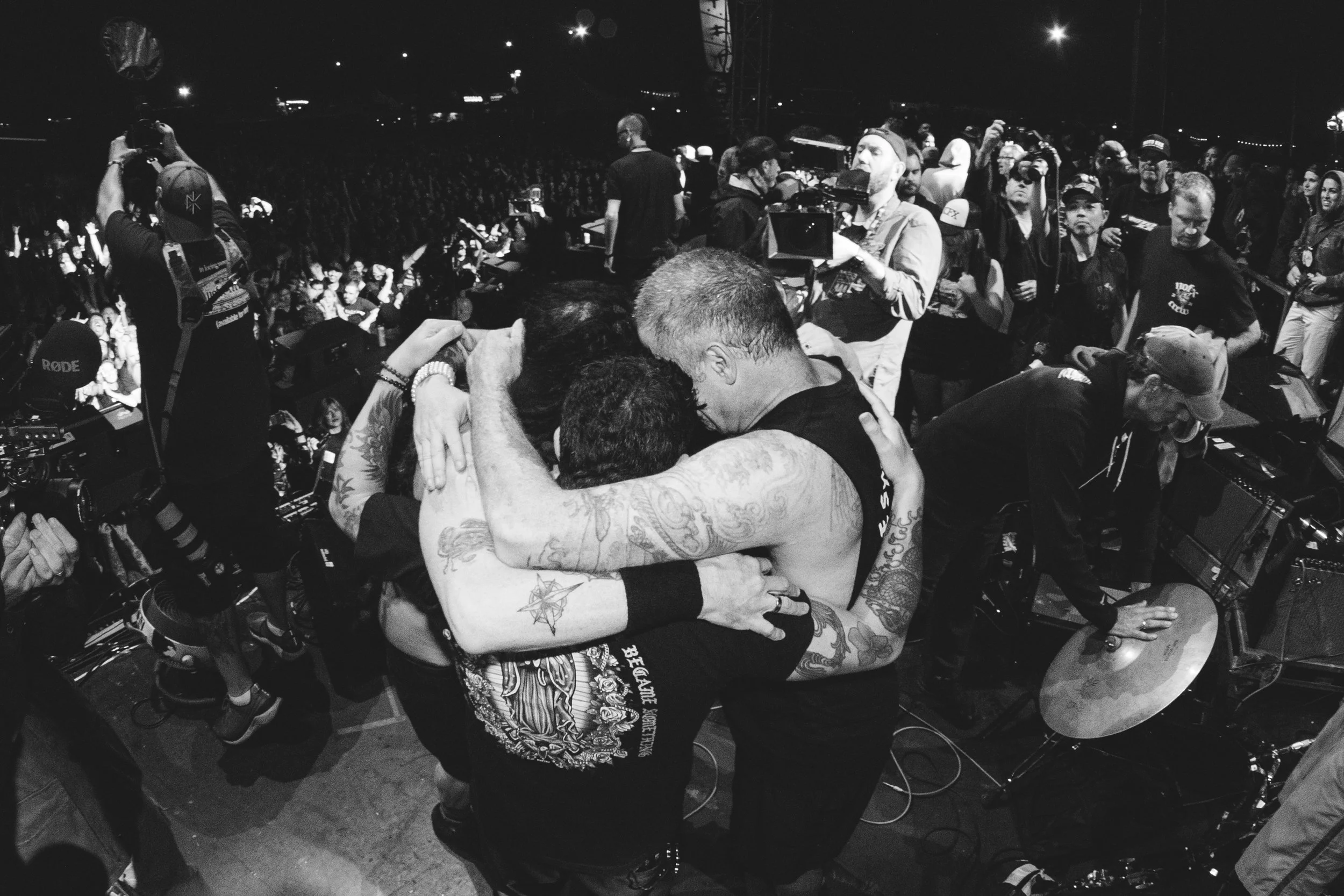 Two people hugging on stage at a concert or music event, surrounded by a crowd of attendees, photographers, and a DJ setup.