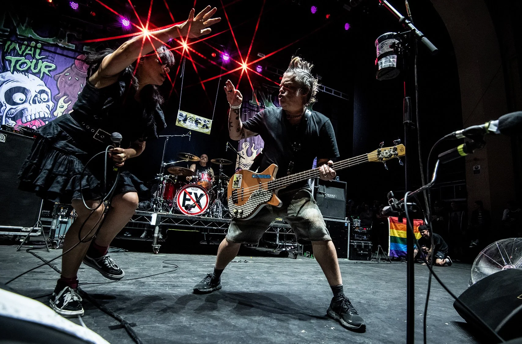 Two musicians perform on stage during a concert, one singing with a microphone and the other playing a bass guitar. A drummer is in the background, and stage lighting features red and purple lights. There is a rainbow pride flag on the side.