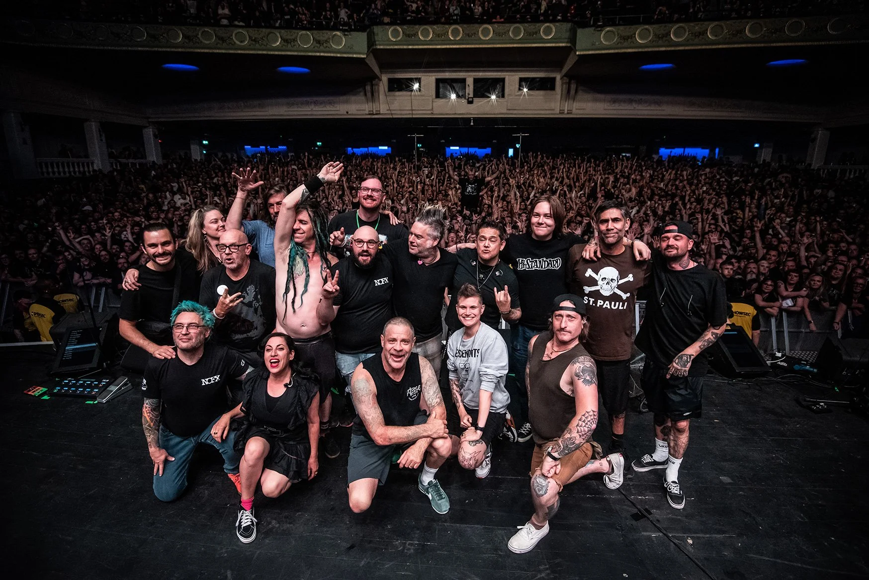 Group of band members and fans on stage at a concert with large crowd in the background.
