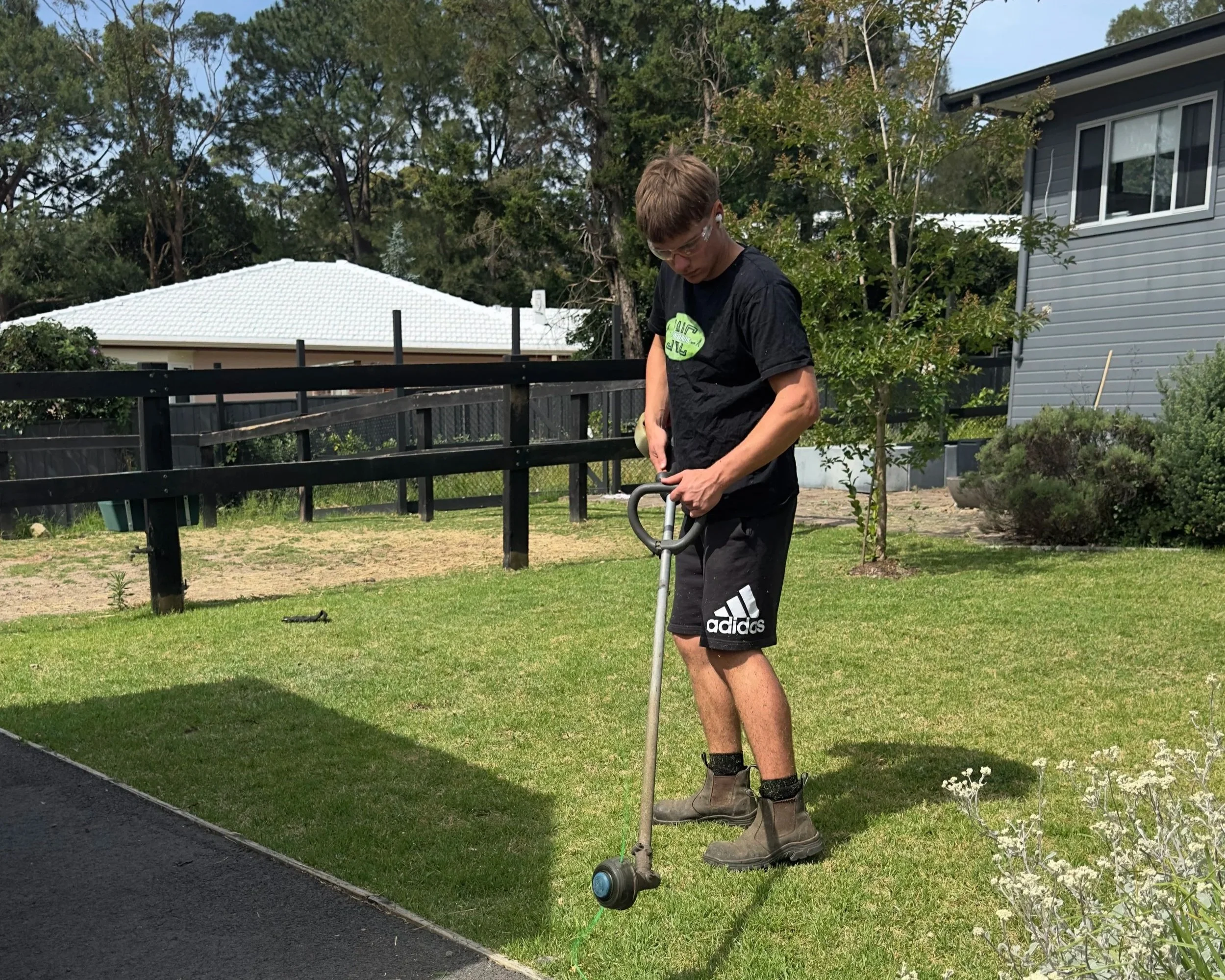 A young man using a grass trimmer to trim the edge of a lawn in a suburban backyard wearing glasses, a black t-shirt, black shorts, and work boots.