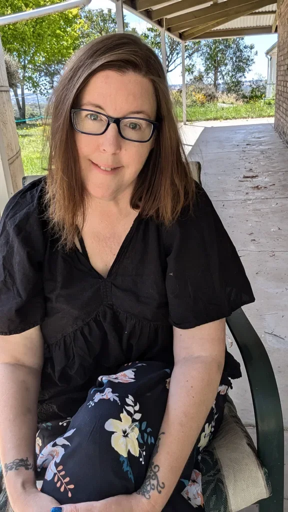 A woman with glasses and brown hair sitting outdoors on a porch, smiling at the camera, with trees and a garden in the background.