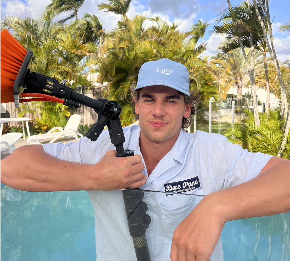 A young man in a uniform shirt and a light blue cap holding a pressure washer wand, smiling, with a backyard pool, palm trees, and a partly cloudy sky in the background.