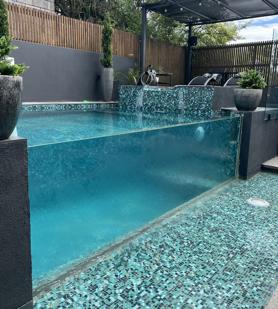 A modern backyard pool with a glass wall, mosaic tiling, and potted plants. There is a shaded area with lounge chairs and garden tools in the background.