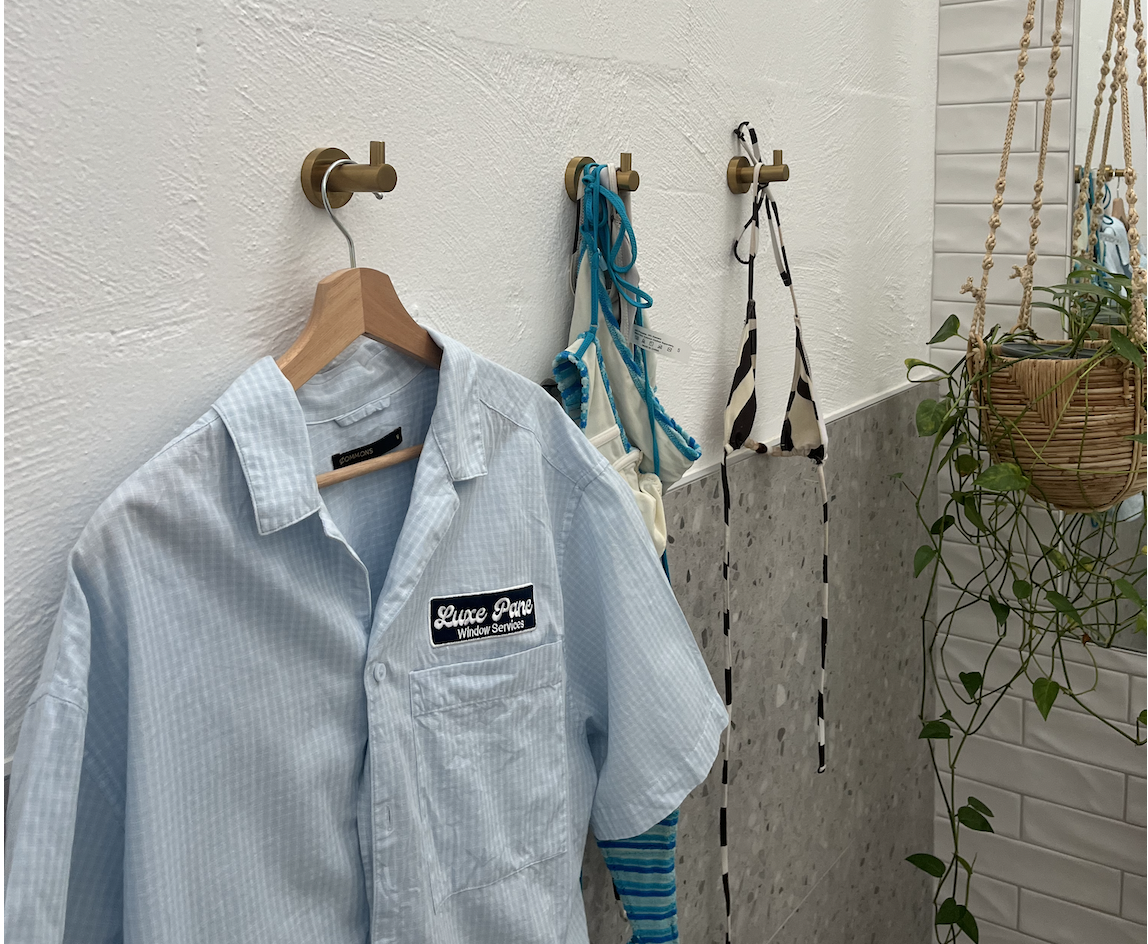 A white shirt with a black patch reading 'Luxe Pane Window Services' hanging on a wooden hanger, attached to a wall with three hooks. Two bras are hanging on the hooks and a plant in a hanging basket is on a shelf to the right.