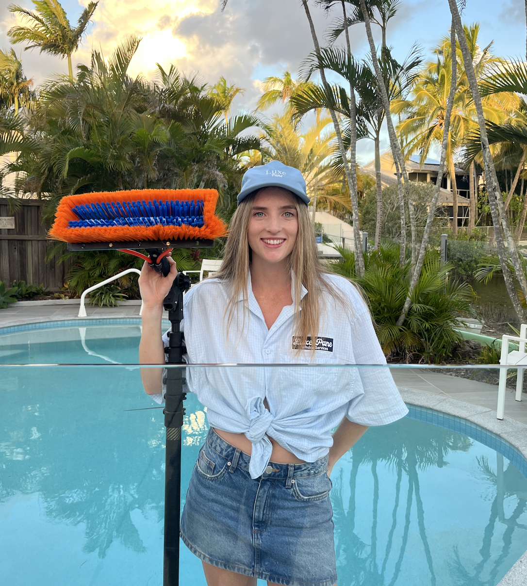A young woman with long blonde hair, wearing a light blue cap and a tied-up white shirt, is standing by a swimming pool holding a leaf blower with an orange and blue brush attachment. She is smiling and the background features tropical palm trees and