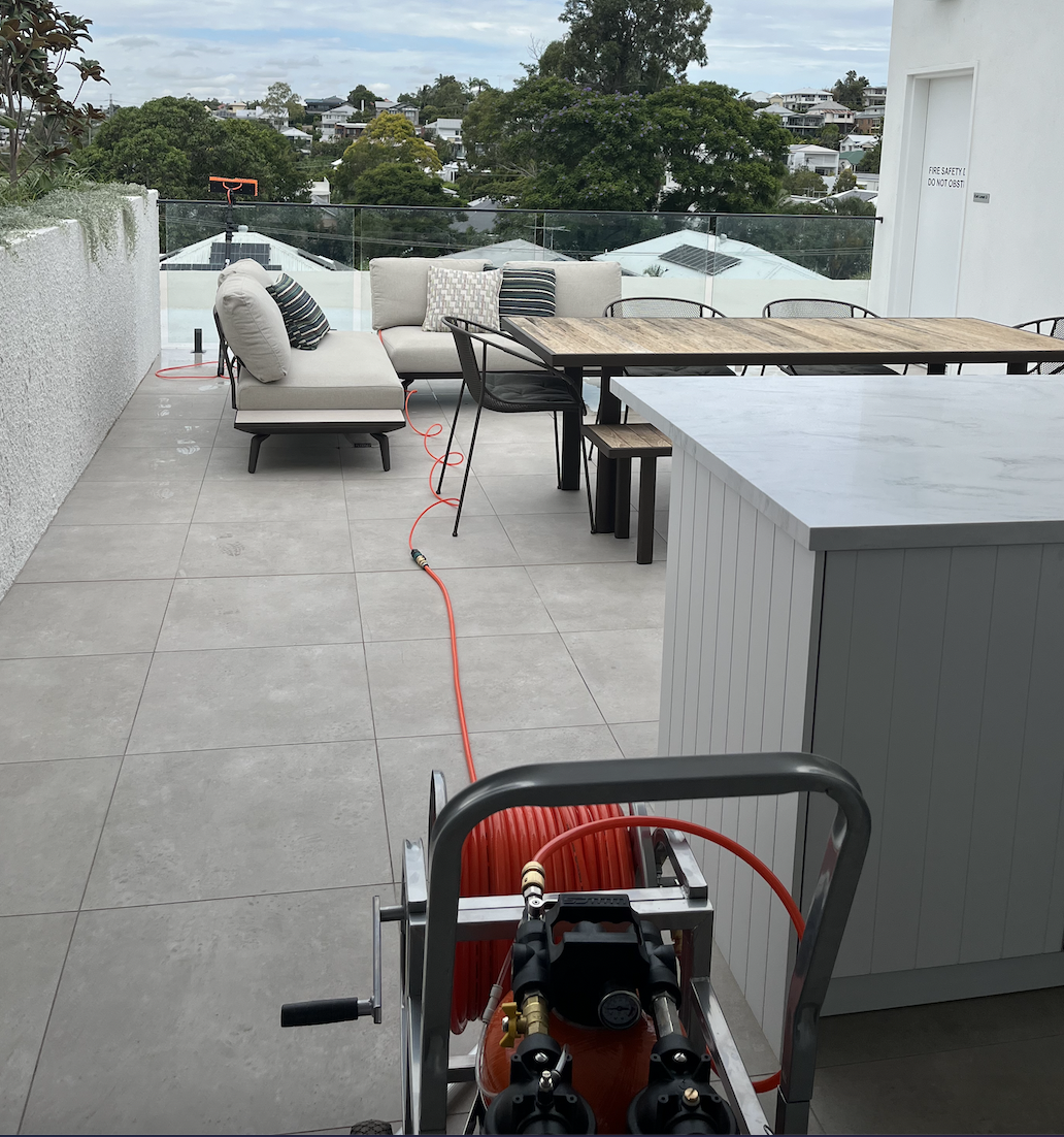 Outdoor balcony with patio furniture, including a sofa with pillows and a wooden dining table with chairs, view of trees and houses in the background, and equipment on the floor connected to a hose.