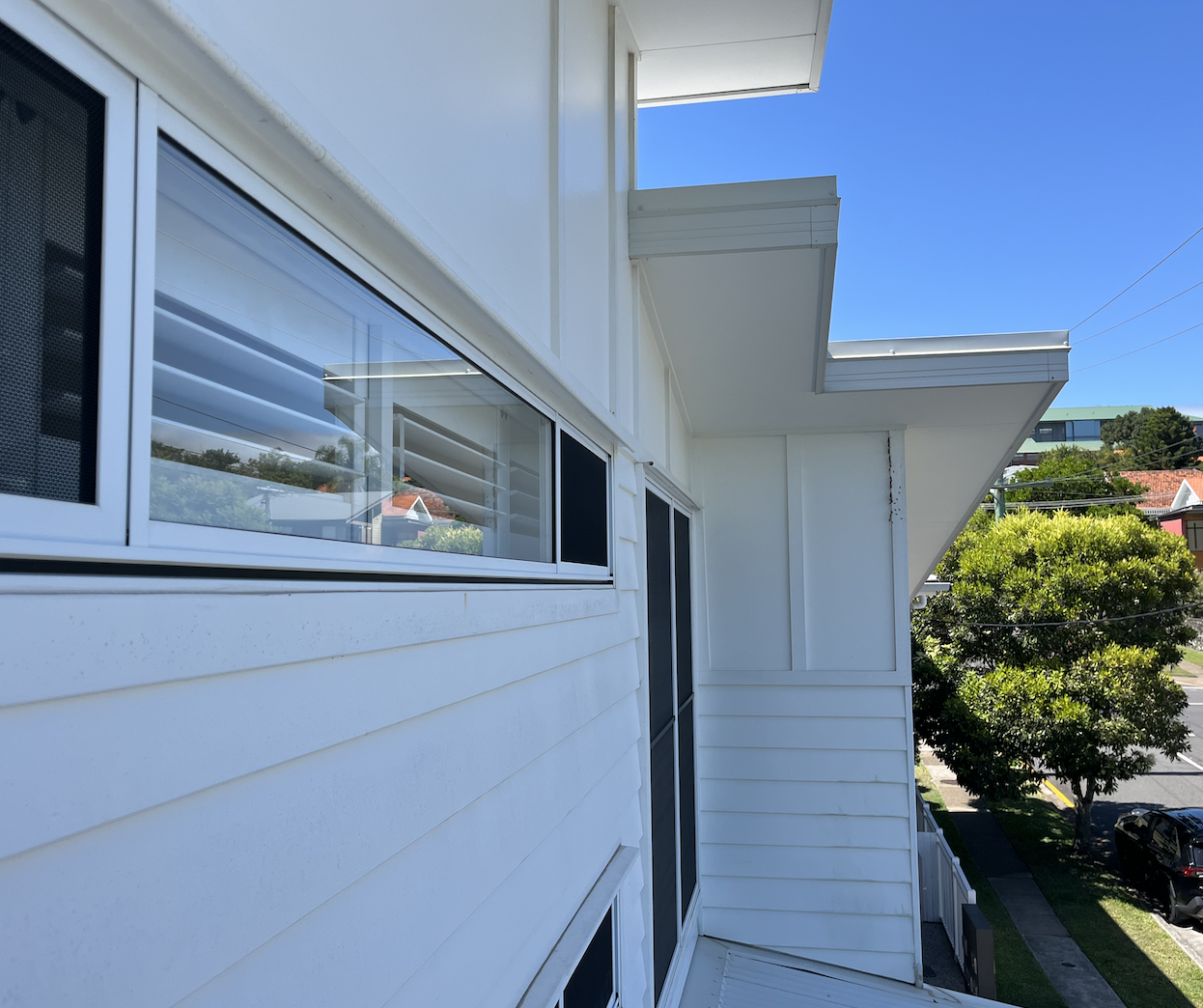 Side view of a white building with horizontal siding, several large windows, and a small balcony with steps, with trees and a street with cars in the background under a clear blue sky.