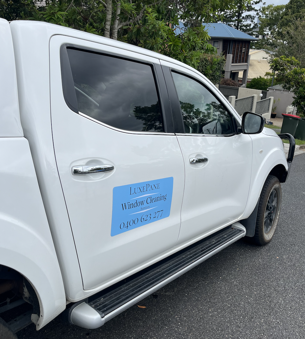 White pickup truck with a blue sign on the door advertising LuxePANE window cleaning services and phone number, parked on a residential street with trees and houses in the background.