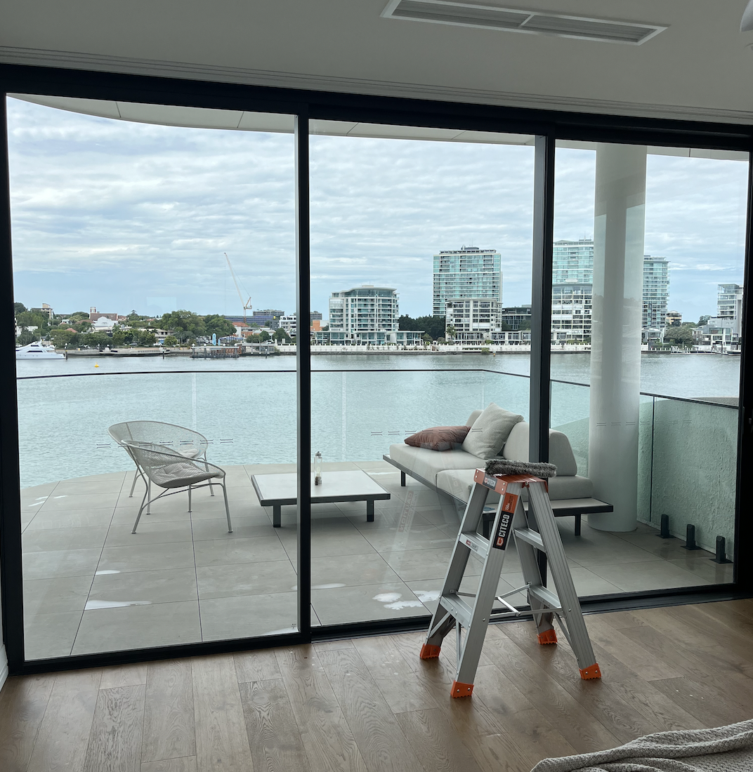 Living room with sliding glass door leading to balcony overlooking water and city skyline, with outdoor seating and a ladder inside.