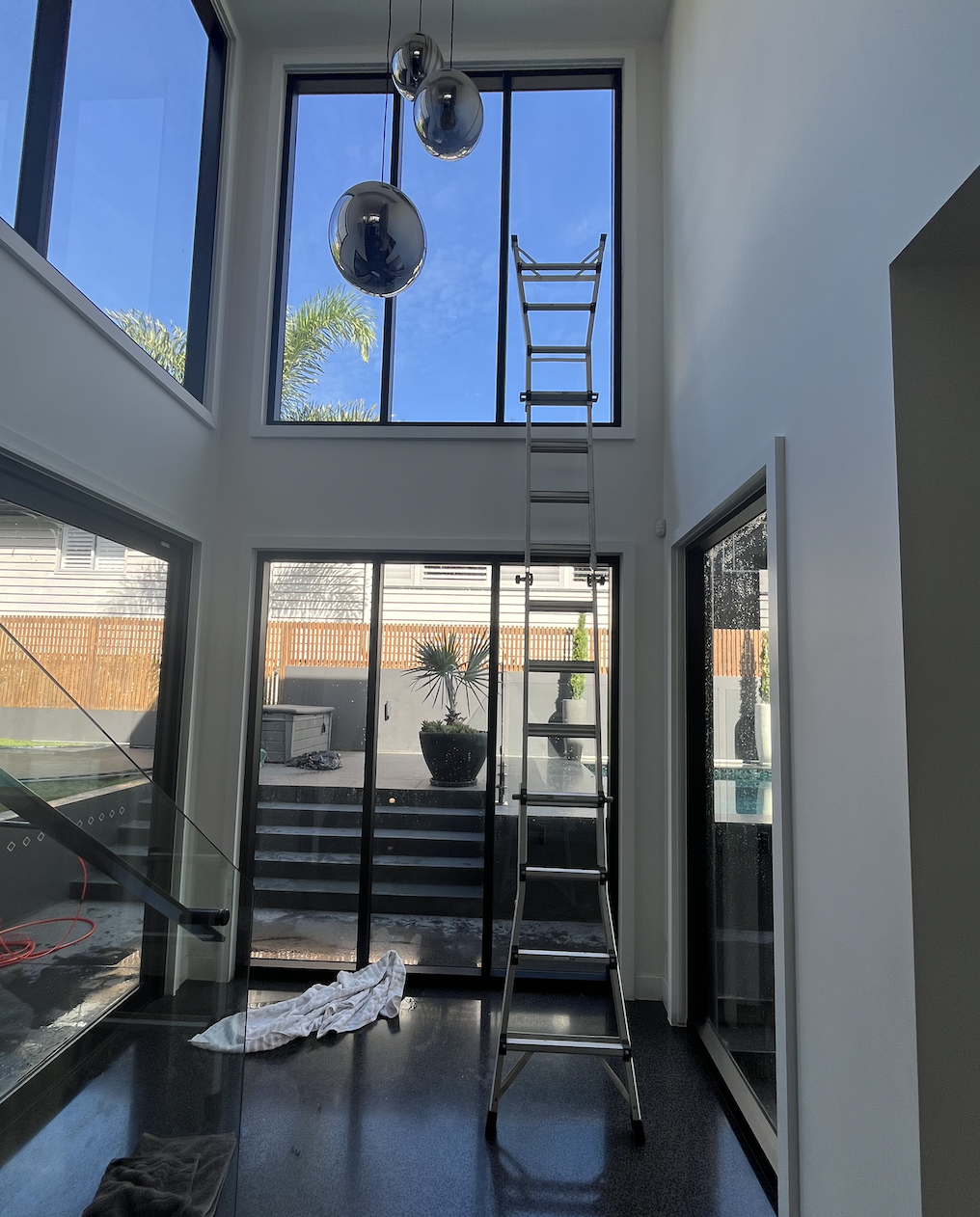 View of a modern interior with high ceilings and large windows, a ladder reaching the upper window, hanging metallic balloons, and a sliding door leading to an outdoor patio with steps and potted plants.