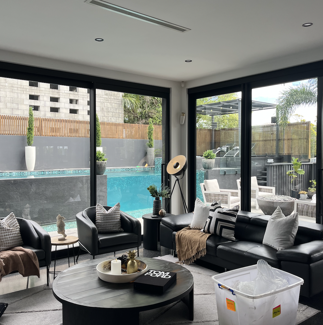 Modern living room with black leather sofas, decorative pillows, and a round coffee table with candles and a pineapple-shaped decor piece, overlooking a pool area through large sliding glass doors.