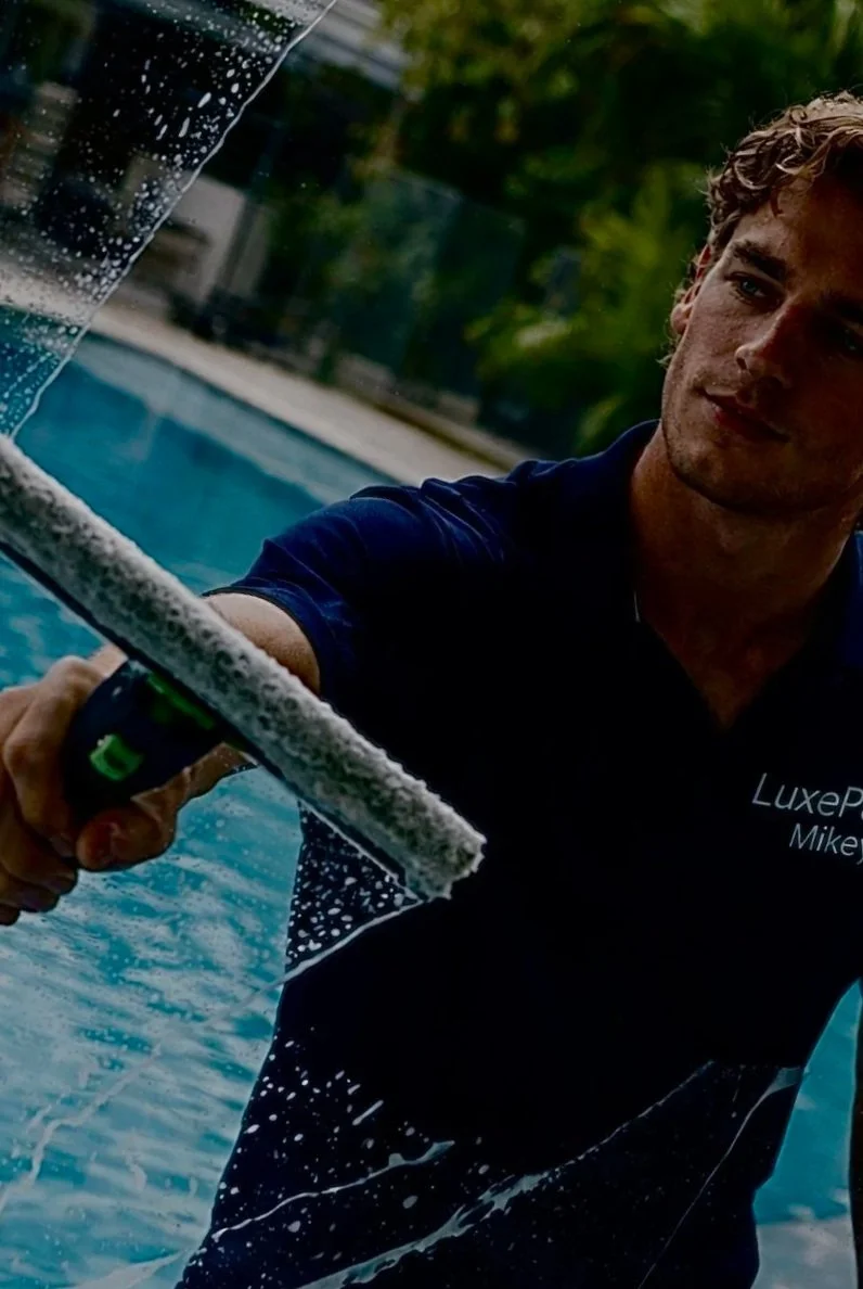 A man cleaning a large glass window or door with a squeegee near a swimming pool, with trees and outdoor scenery in the background.