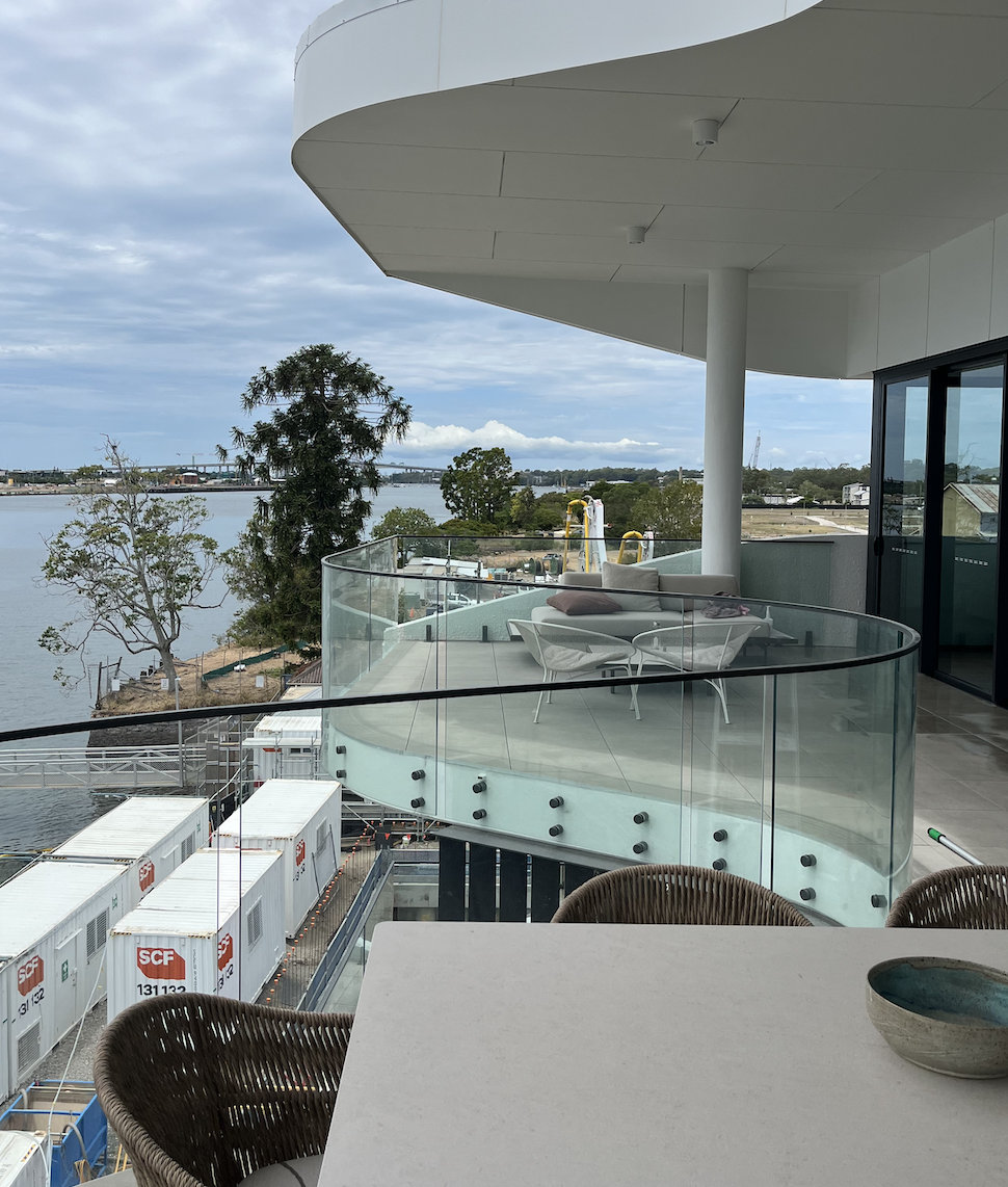 View from a balcony overlooking a river, with outdoor seating, trees, and construction containers nearby.
