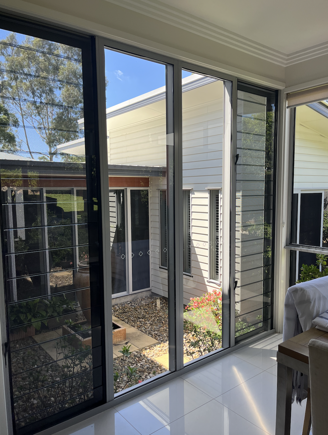 Interior view of a room with large glass sliding doors and windows, opening to a small outdoor garden with rocks, plants, and a neighboring white house.