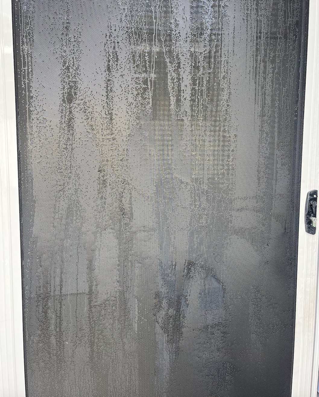 A close-up of a window screen with water streaks and smudges, framed by a white window frame.