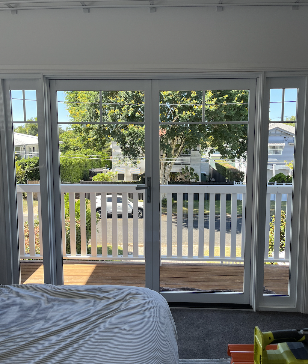 View of a porch through a sliding glass door, with a white railing, trees, houses, and a parked car outside, from inside a bedroom with a bed in the lower left corner and a yellow tool on the floor.