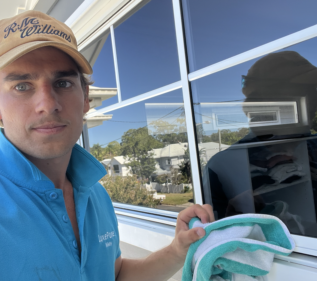 Man cleaning a window with a cloth, wearing a blue shirt and a beige cap.