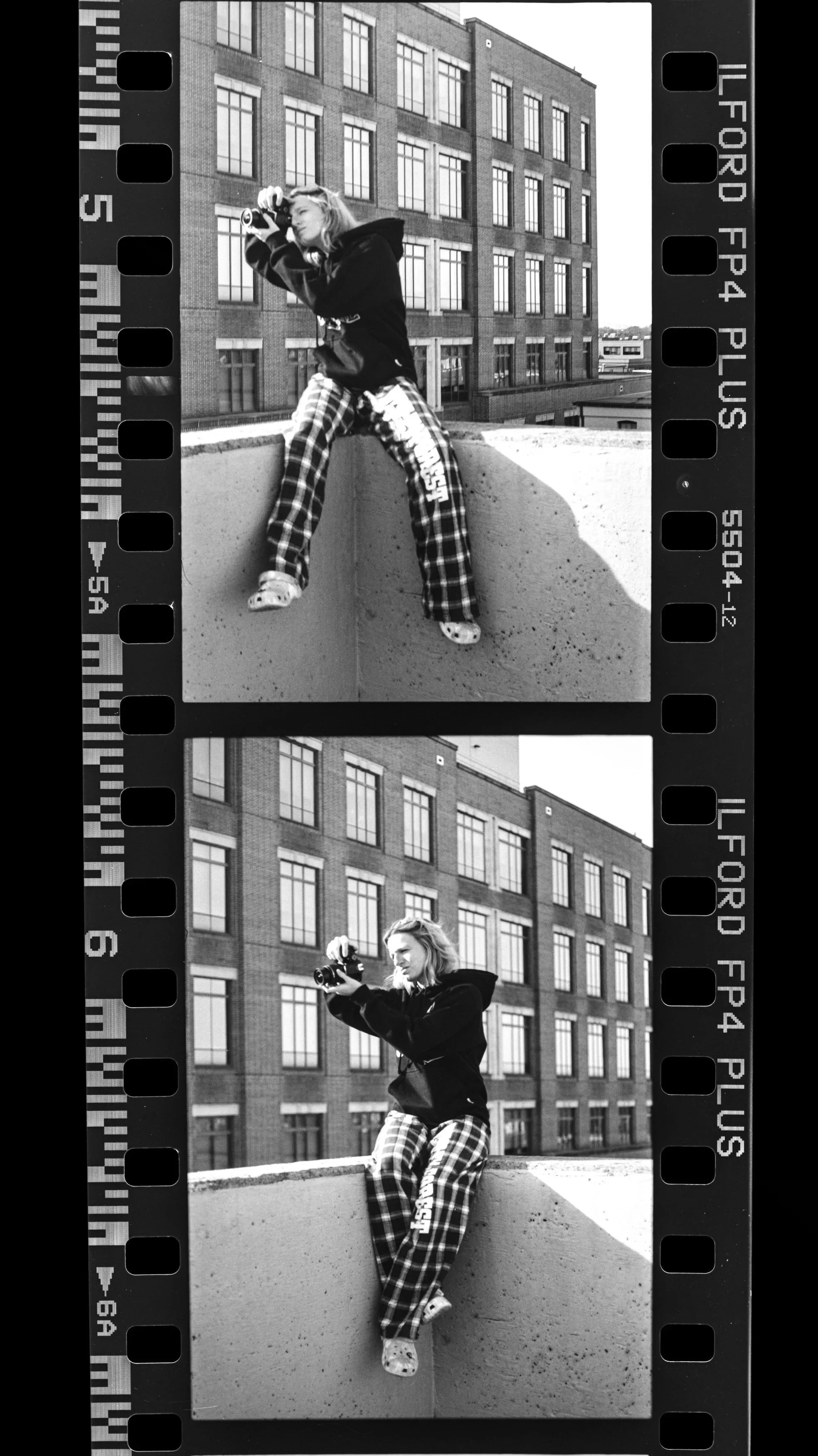 Black and white photograph of a woman on a rooftop, taking pictures with a camera. She is sitting on the edge of the rooftop, wearing a hoodie, patterned pants, and Crocs, with a building in the background.