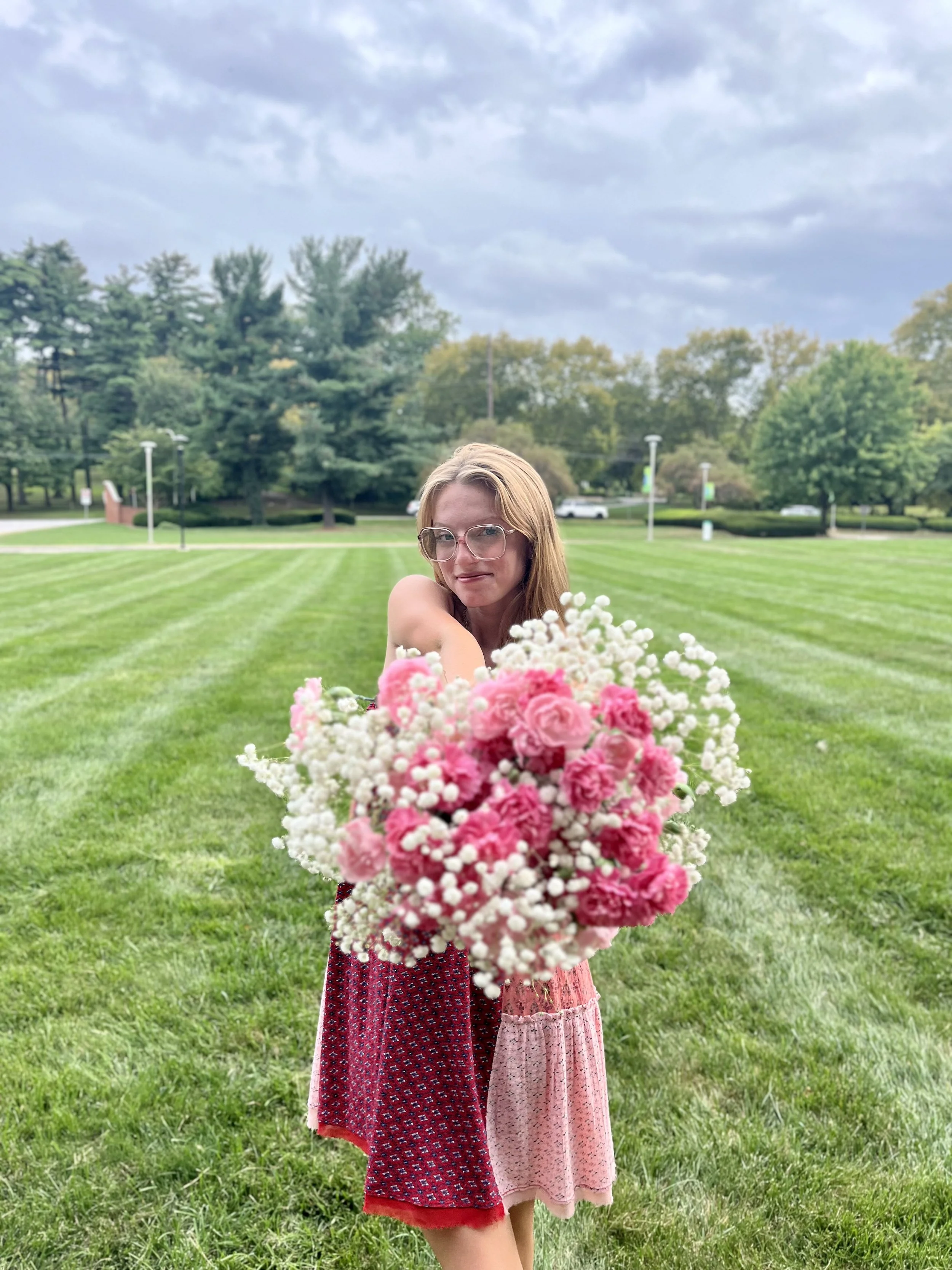 A young woman with glasses holding a large bouquet of pink and white flowers outdoors on a grassy field with trees and overcast sky in the background.