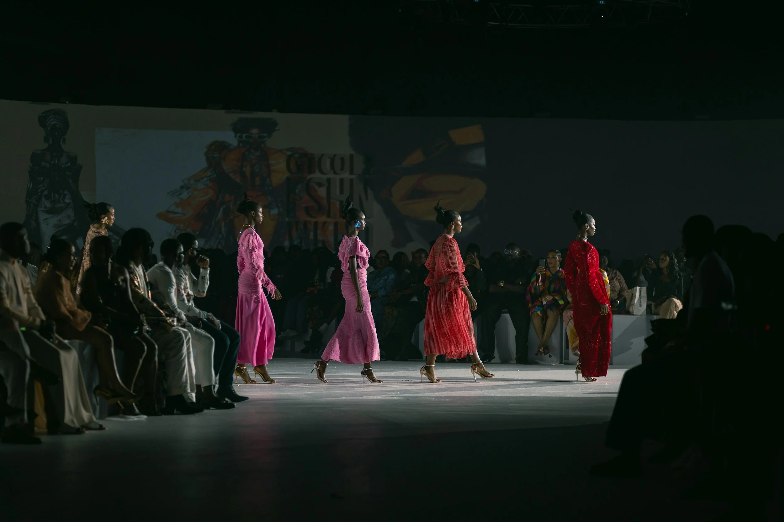 Models walking down runway at a fashion show, with seated audience on both sides and a large screen displaying fashion graphics in the background.