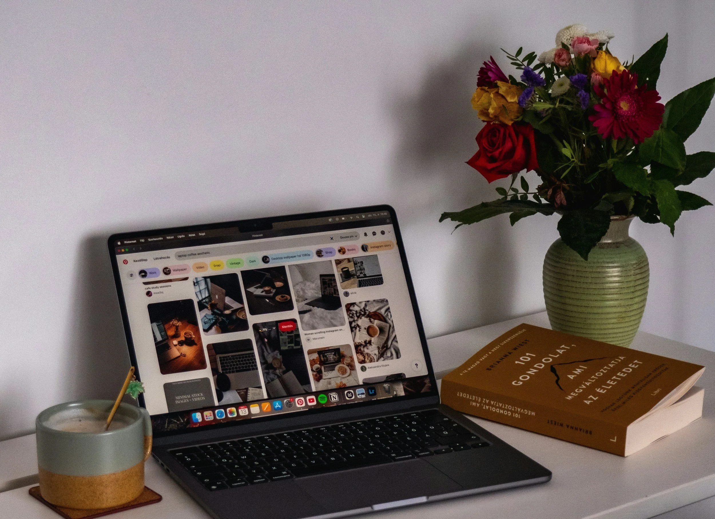 A desk setup with an open laptop displaying a Pinterest board, a large book titled '101 GONDOLA', a potted flower arrangement, and a cup with a green stir stick on a coaster.