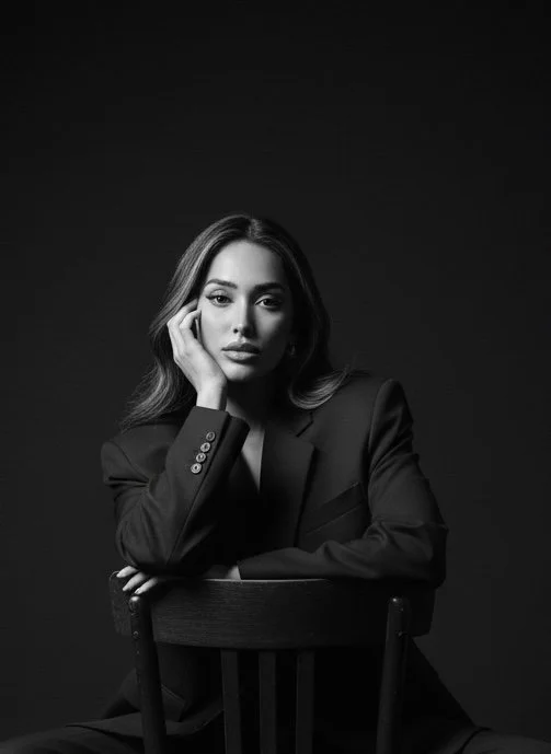 Black and white portrait of a woman in a suit, seated on a chair, resting her head on her hand, with a serious expression.
