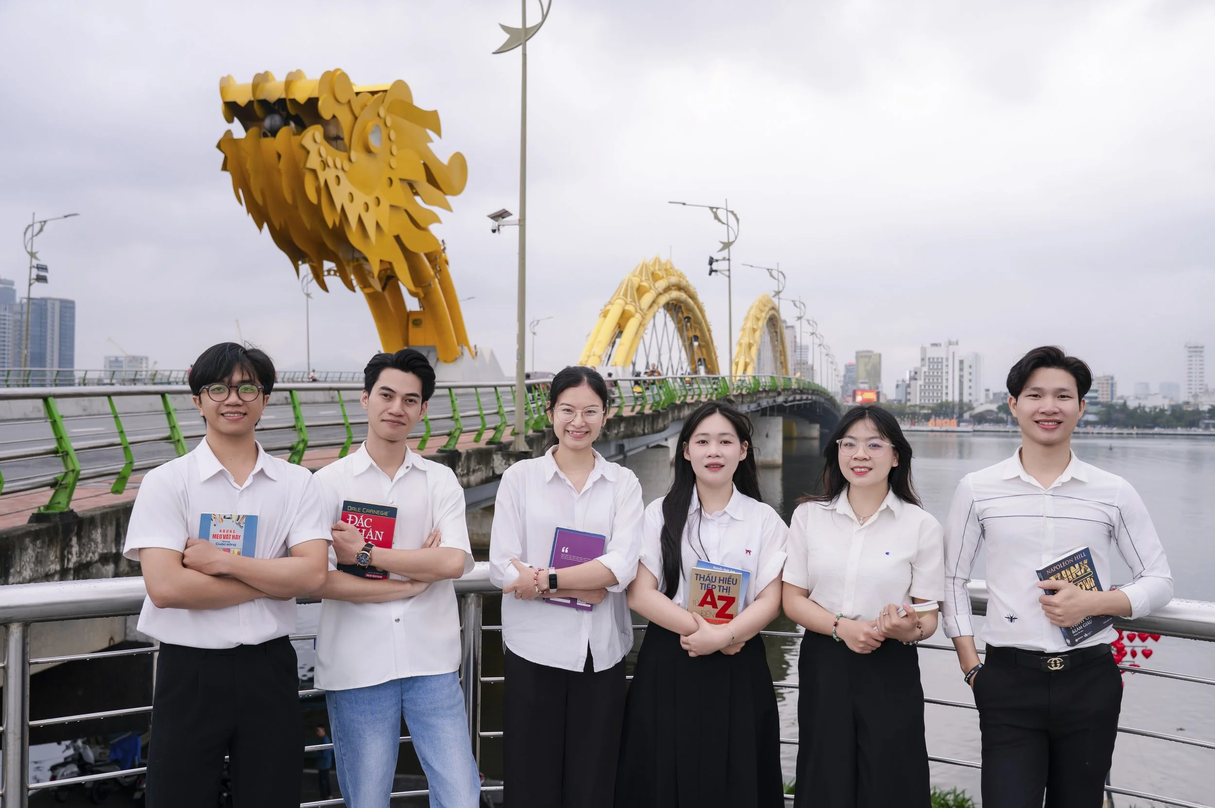 Sechs junge Menschen stehen an einer Flusspromenade und halten Bücher in den Armen, im Hintergrund eine Brücke mit goldfarbenen Drachenkopf-Architektur und einer Stadt mit Hochhäusern bei bewölktem Himmel.