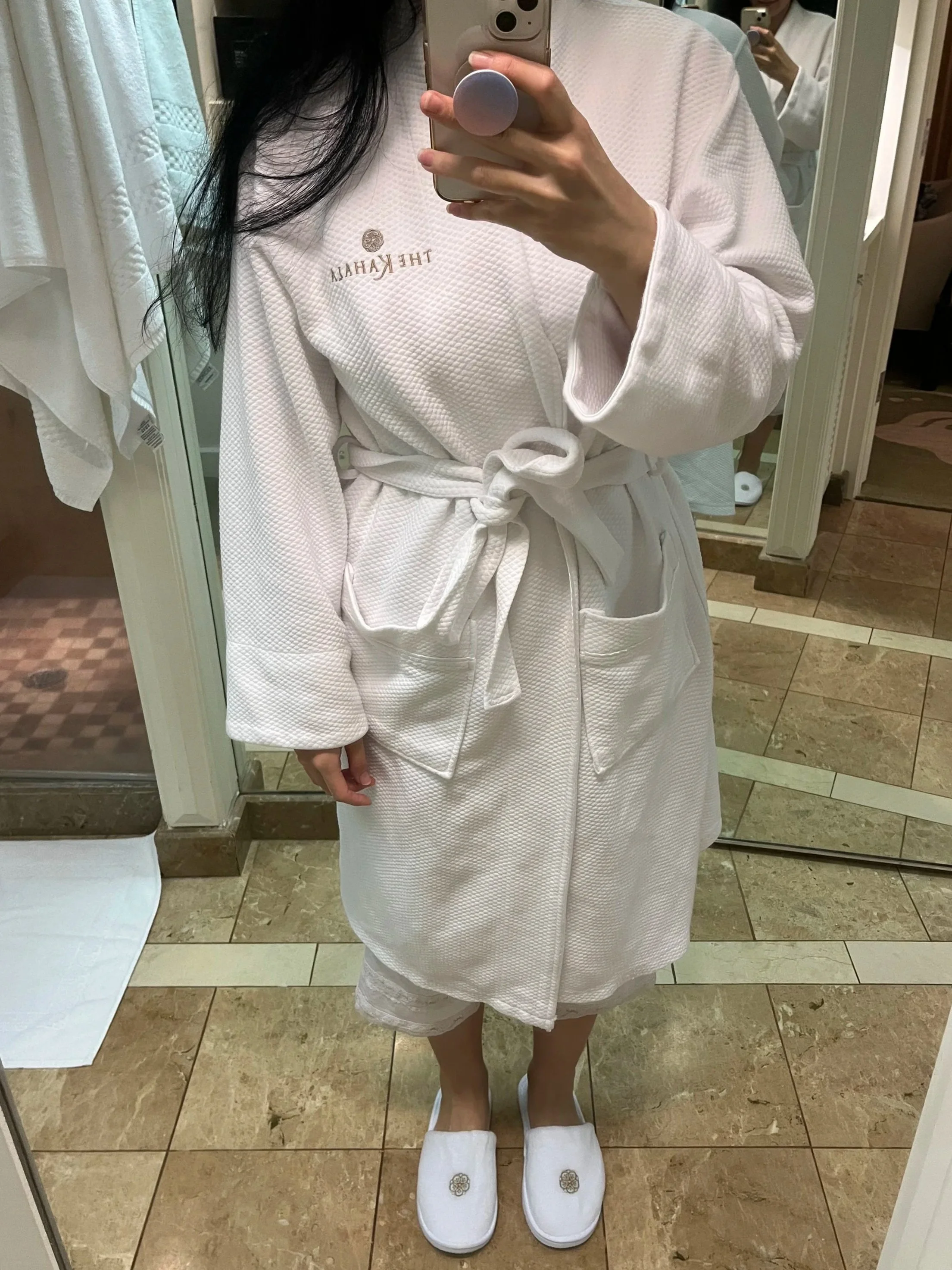a woman taking a full body selfie in hotel mirror wearing a white robe and white slippers