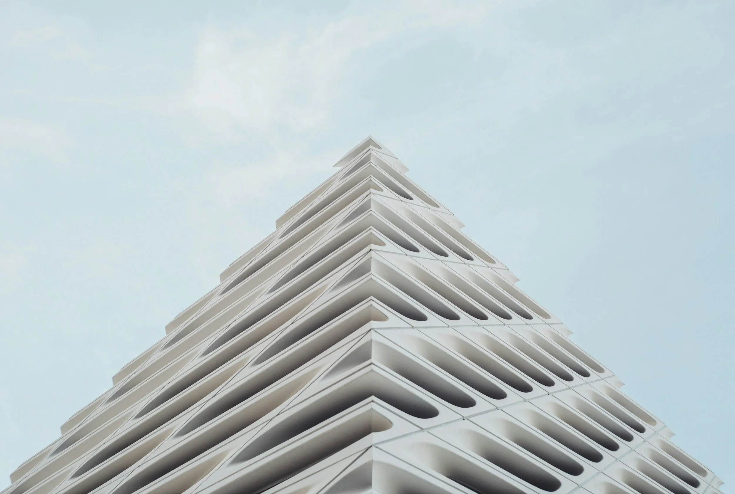 A modern multi-story building with a white facade, viewed from below against a cloudy sky. The building features geometric design with repetitive cut-out patterns on each floor.