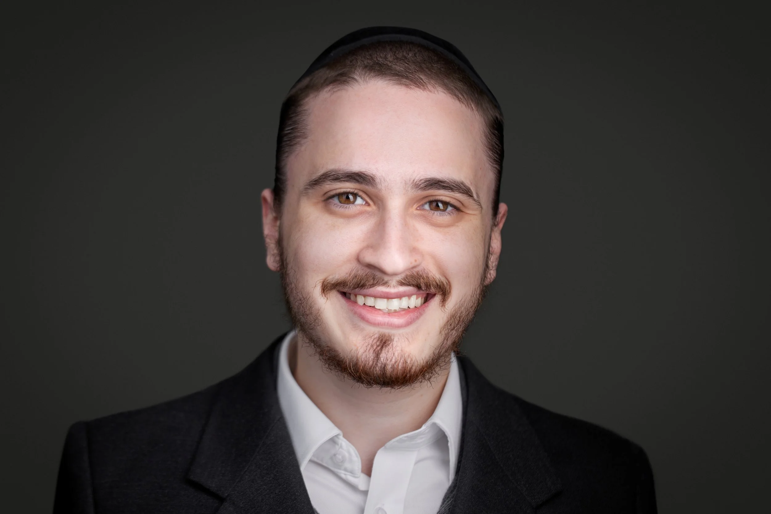 A smiling young man with short brown hair, light brown eyes, and a beard, wearing a white shirt and black blazer in front of a dark background.