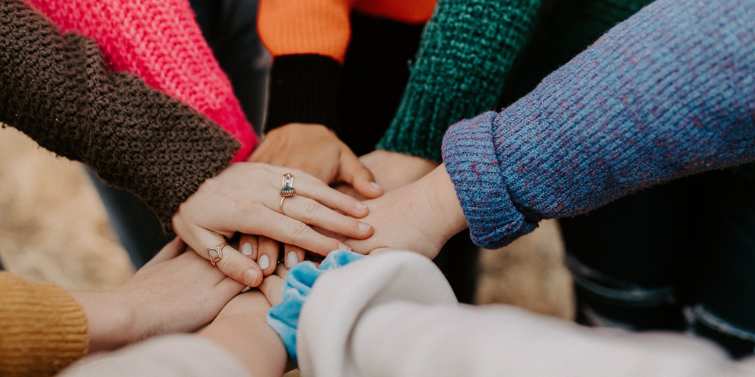 Group of people stacking their hands together in a show of unity or teamwork, wearing colorful sweaters and rings.
