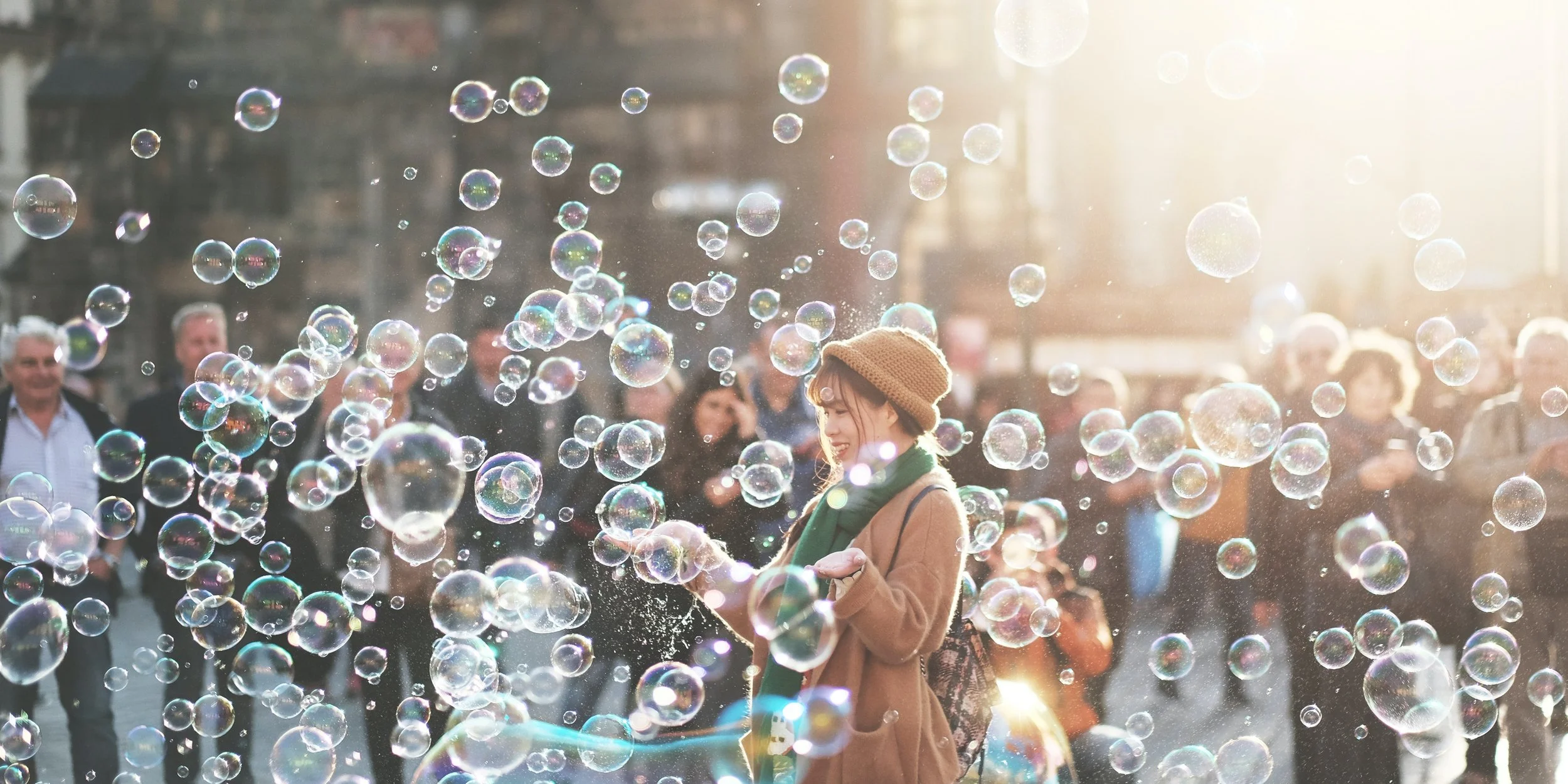 A young woman in a brown coat and knit hat is smiling and playing with soap bubbles in a busy outdoor city square during daylight, with sunlight shining from the background and a crowd of people watching.