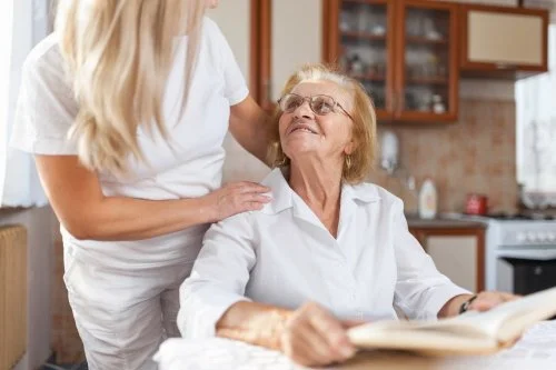 A caregiver standing next to an elderly woman sitting at a kitchen table, smiling and reading a book.