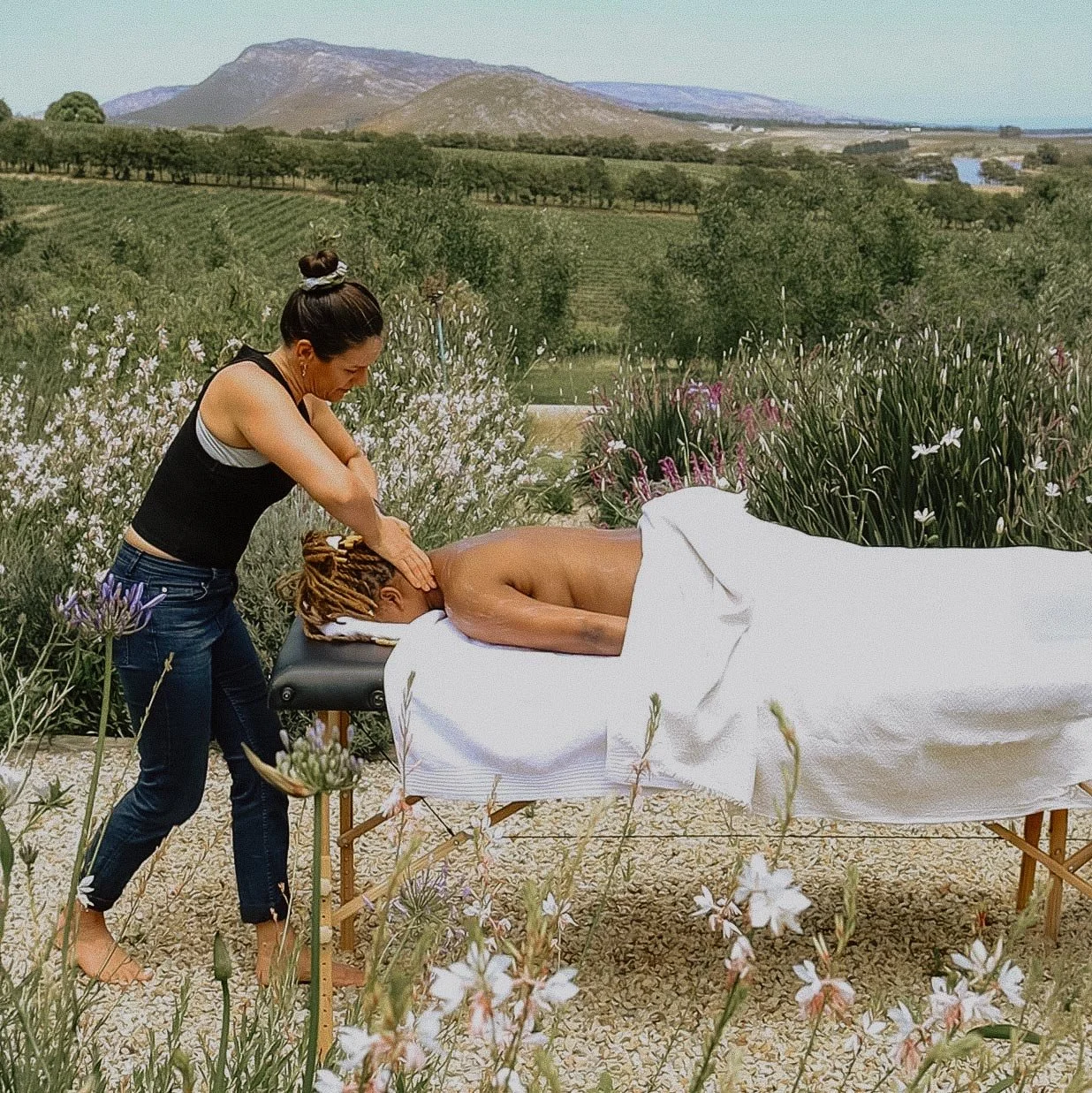 Woman giving a massage outdoors to a man lying face down on a massage table, surrounded by flowering plants and countryside landscape in the background. Private luxury villa (accommodation) in Hemel-en-Aarde, Hermanus.