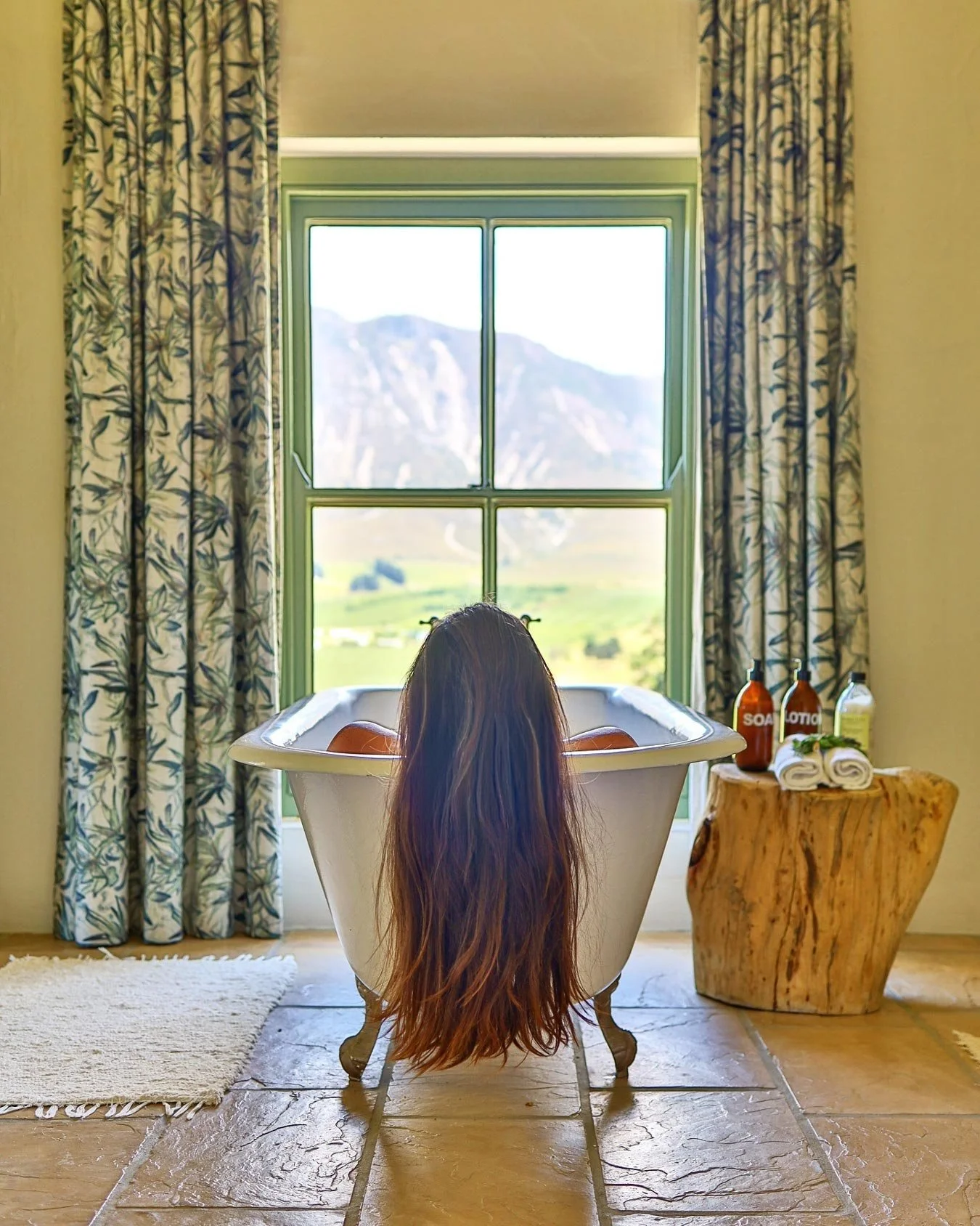 Person with long hair sitting in a vintage bathtub filled with water, facing a large window overlooking mountains, with bath products and towels on a wooden side table by the tub. Private luxury villa (accommodation) in Hemel-en-Aarde, Hermanus.