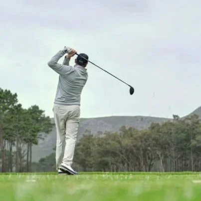 A person in gray attire swinging a golf club on a golf course with green grass, trees, and mountains in the background. Private luxury villa (accommodation) in Hemel-en-Aarde, Hermanus.