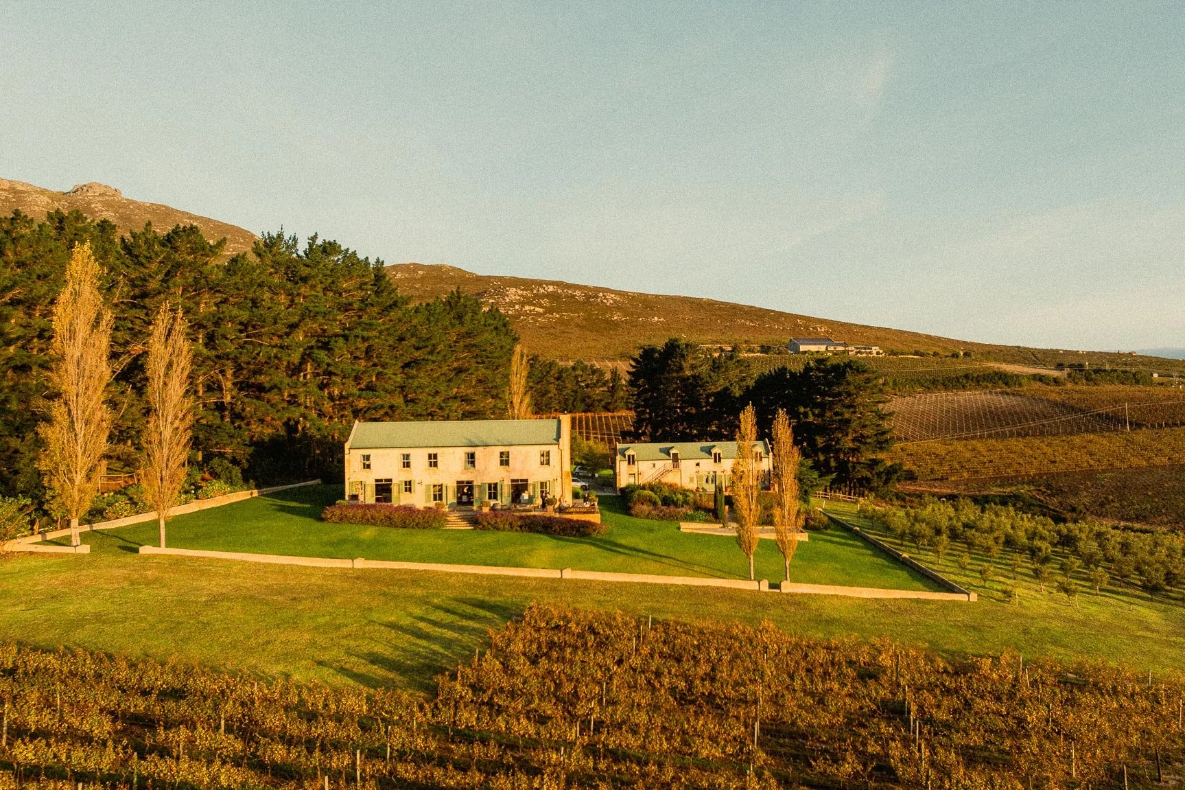 A scenic landscape of a vineyard with a large house and smaller buildings, surrounded by trees, hills, and farmland under a clear sky during sunset. Private luxury villa (accommodation) in Hemel-en-Aarde, Hermanus.