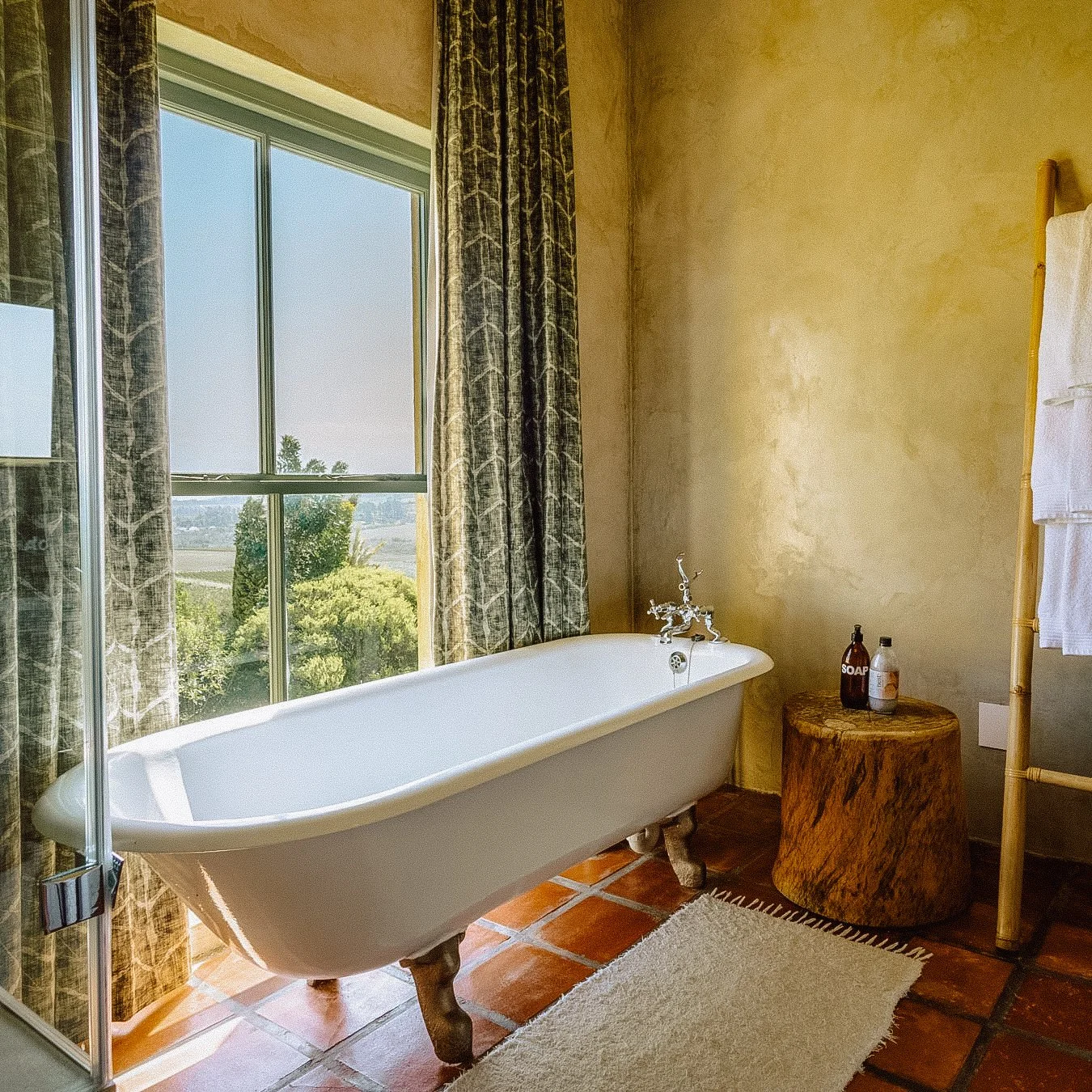 A rustic bathroom features a free-standing clawfoot bathtub next to a large window with patterned curtains, a wooden side table with soap bottles, and a bamboo towel rack with towels. Private luxury villa (accommodation) in Hemel-en-Aarde, Hermanus.