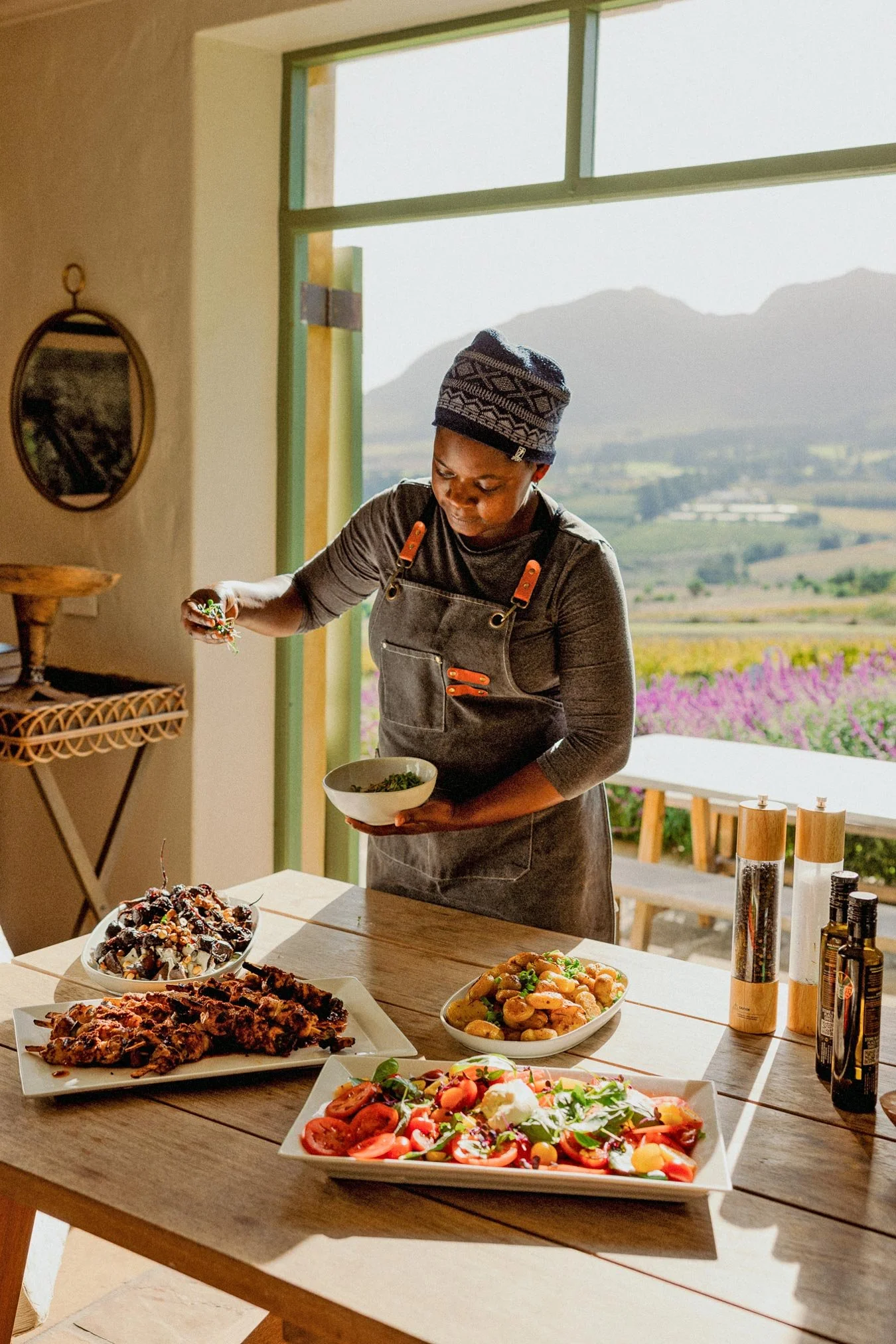 Woman preparing food at a table filled with various dishes, inside a room with open windows revealing mountains and fields outside. Private luxury villa (accommodation) in Hemel-en-Aarde, Hermanus.