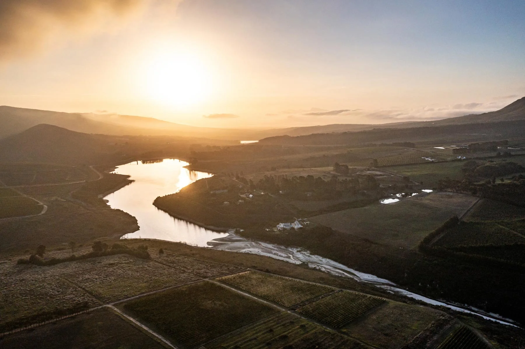 Landscape view of a river winding through fields and farmland with the sun setting in the background, casting a warm glow over the scene. Private luxury villa (accommodation) in Hemel-en-Aarde, Hermanus.